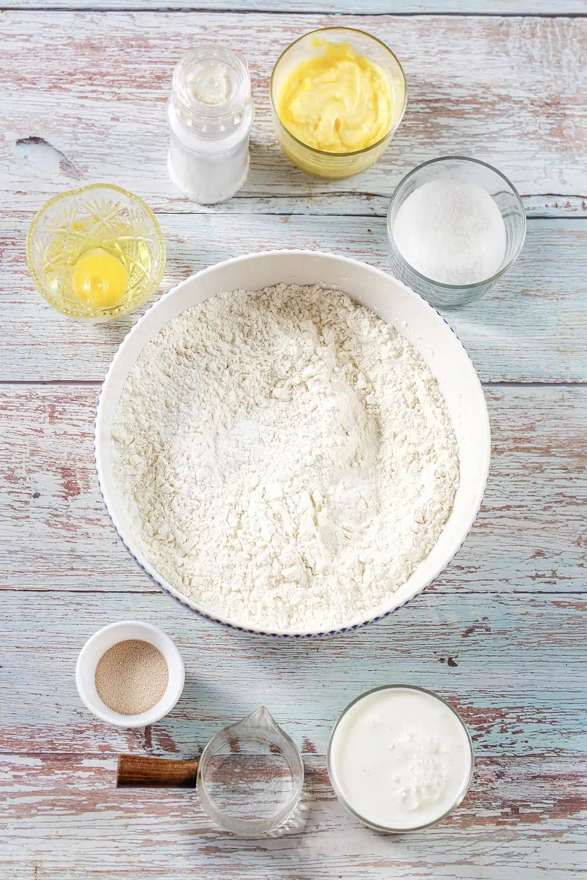 Flour, milk, butter, sugar, an egg and yeast measured into bowls on a painted board