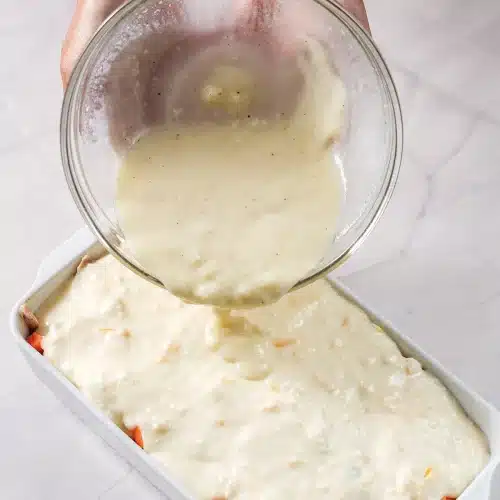 Thick pale batter poured over the meat and vegetable layer in a white dish