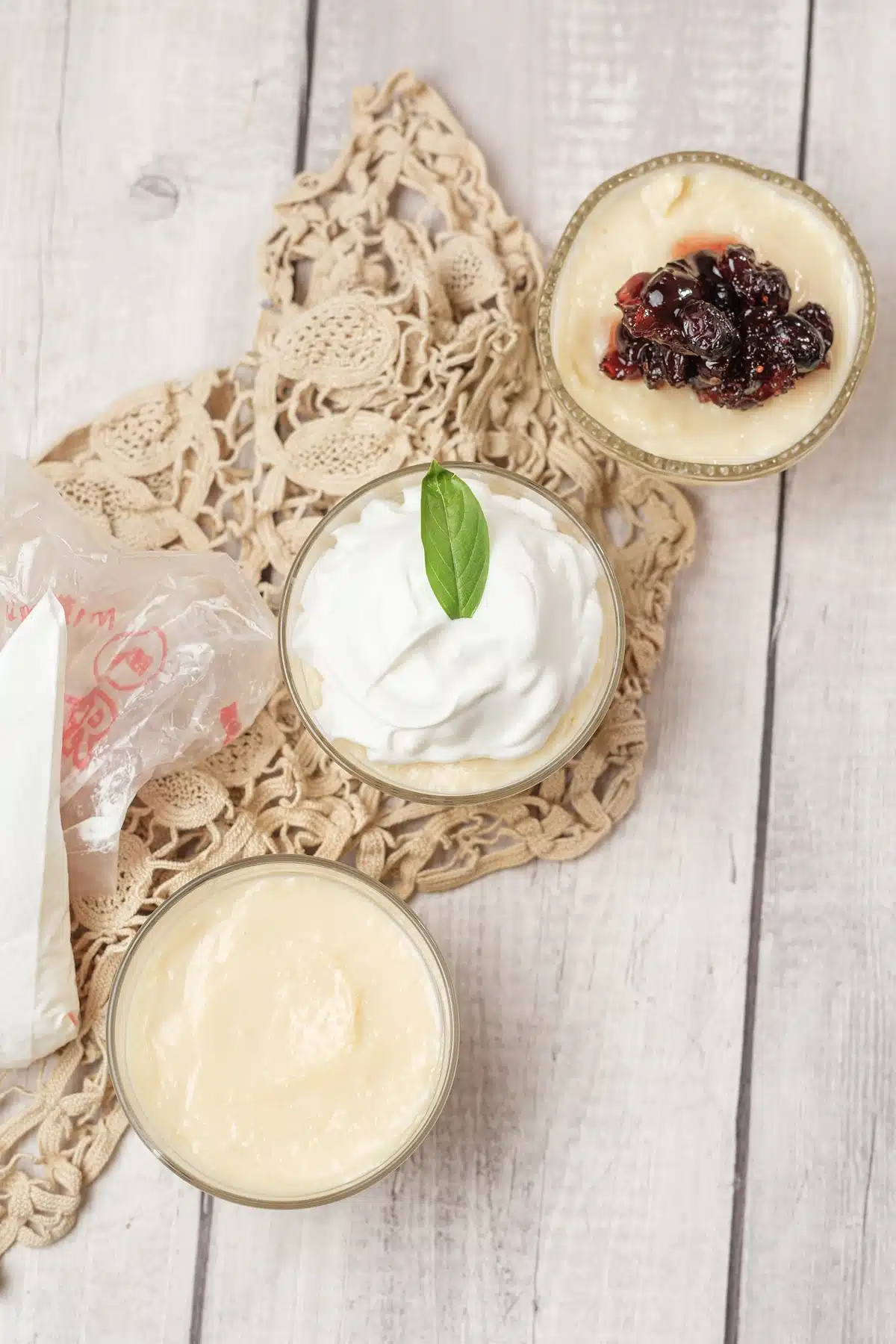 Homemade vanilla pudding in three glasses on a lace doily, one plain, one with whipped cream and one with cranberry compote