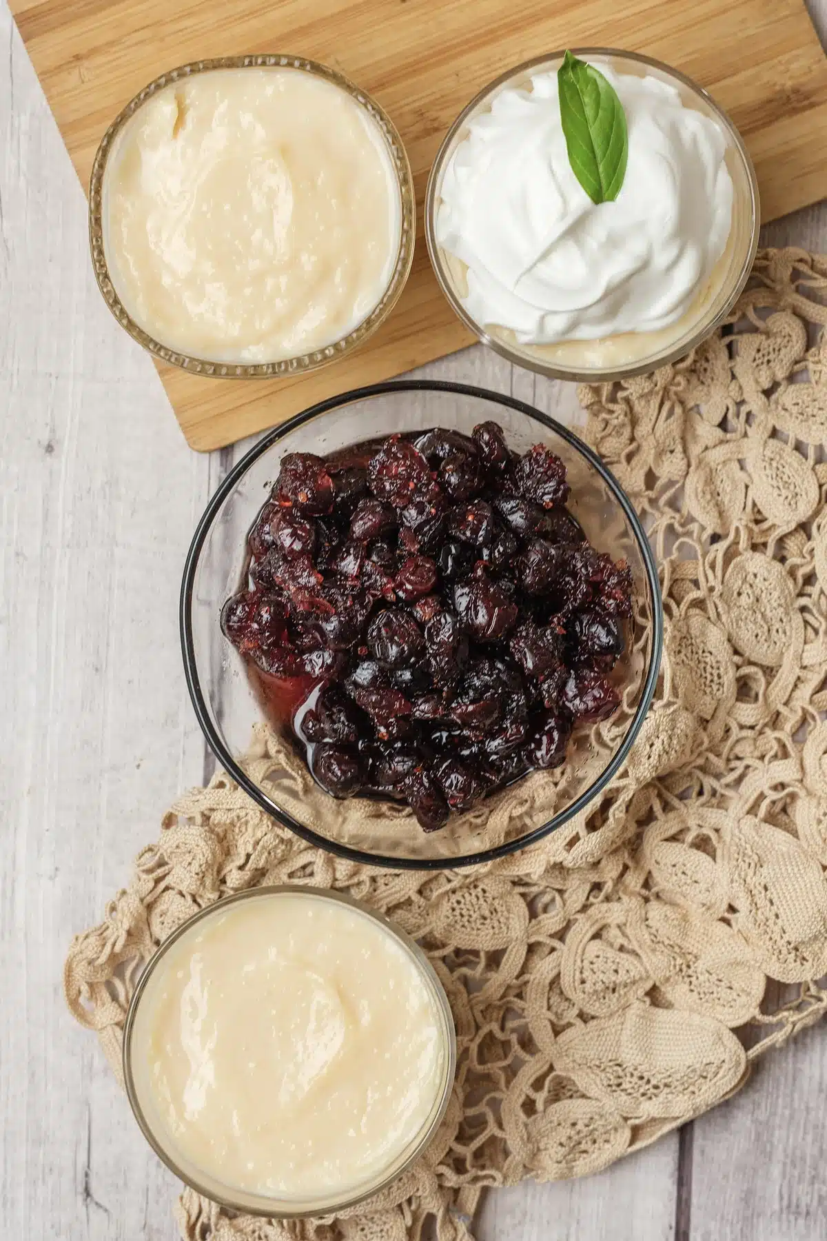 Homemade vanilla pudding in two glasses on a wooden board, one with whipped cream, beside a bowl of cranberry compote