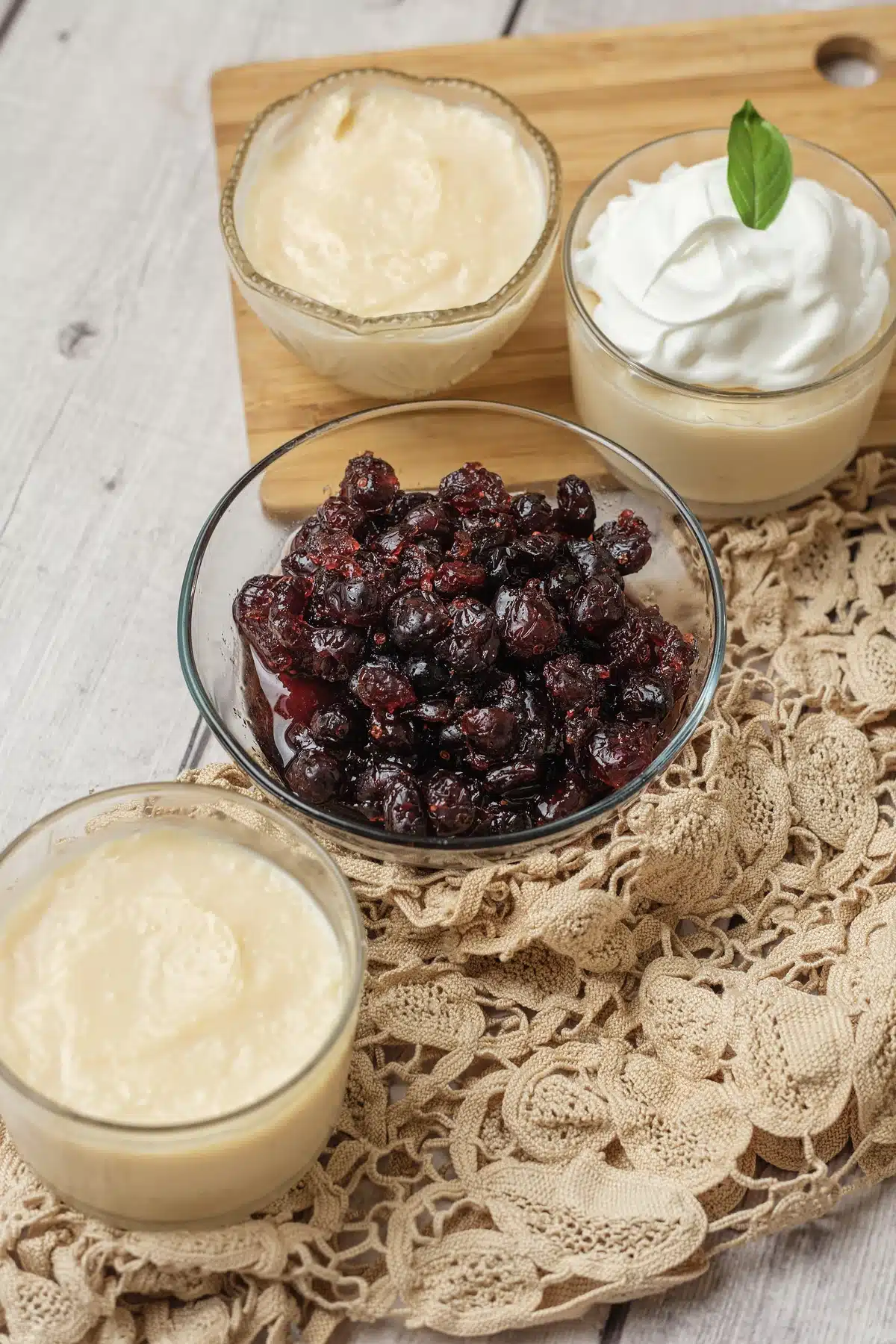 An angled view of two glasses on a board, one topped with whipped cream, and a bowl of dark glossy compote in front