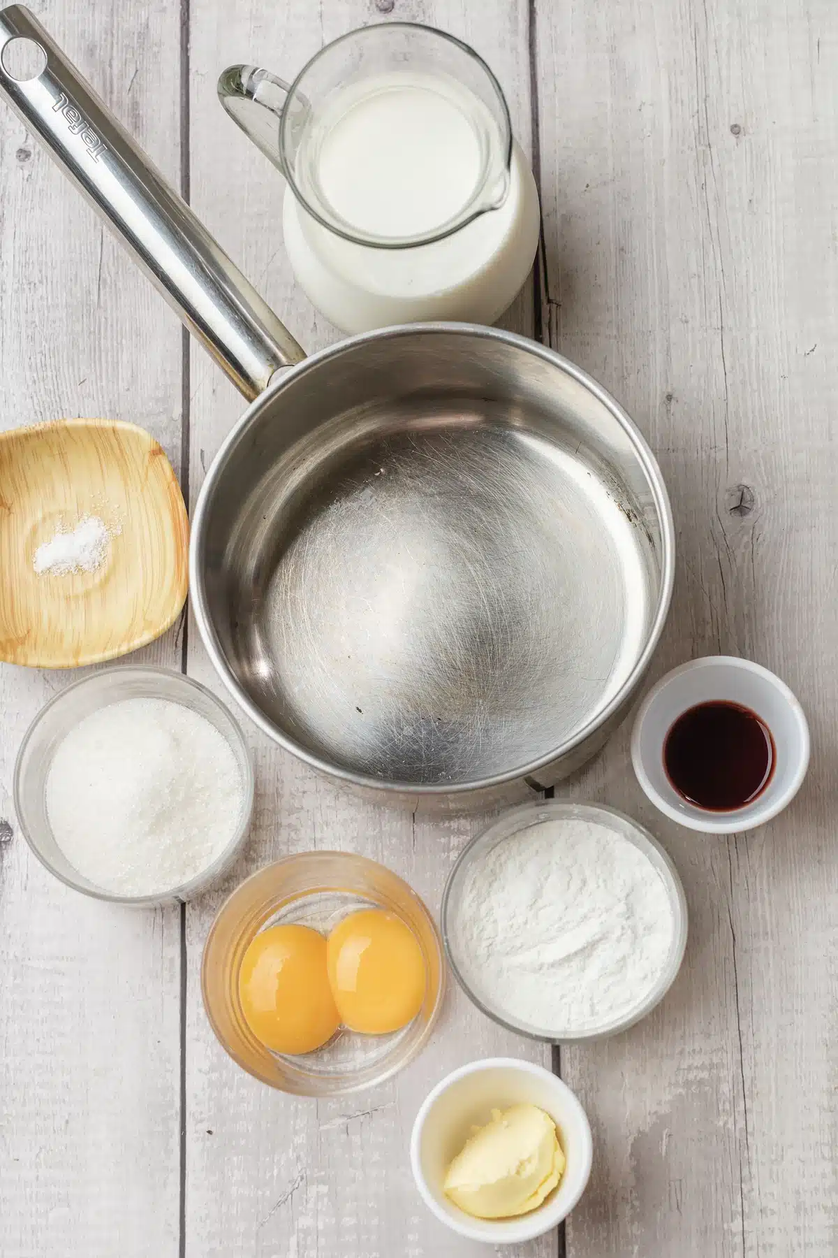 A saucepan with milk, sugar, cornstarch, egg yolks, butter, vanilla and salt set out around it