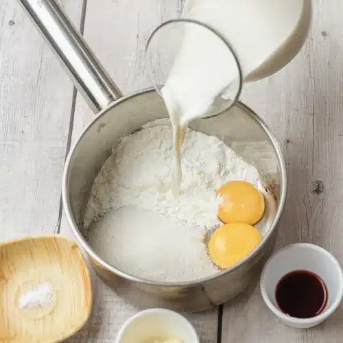 Milk poured from a jug into a saucepan holding sugar, cornstarch and egg yolks
