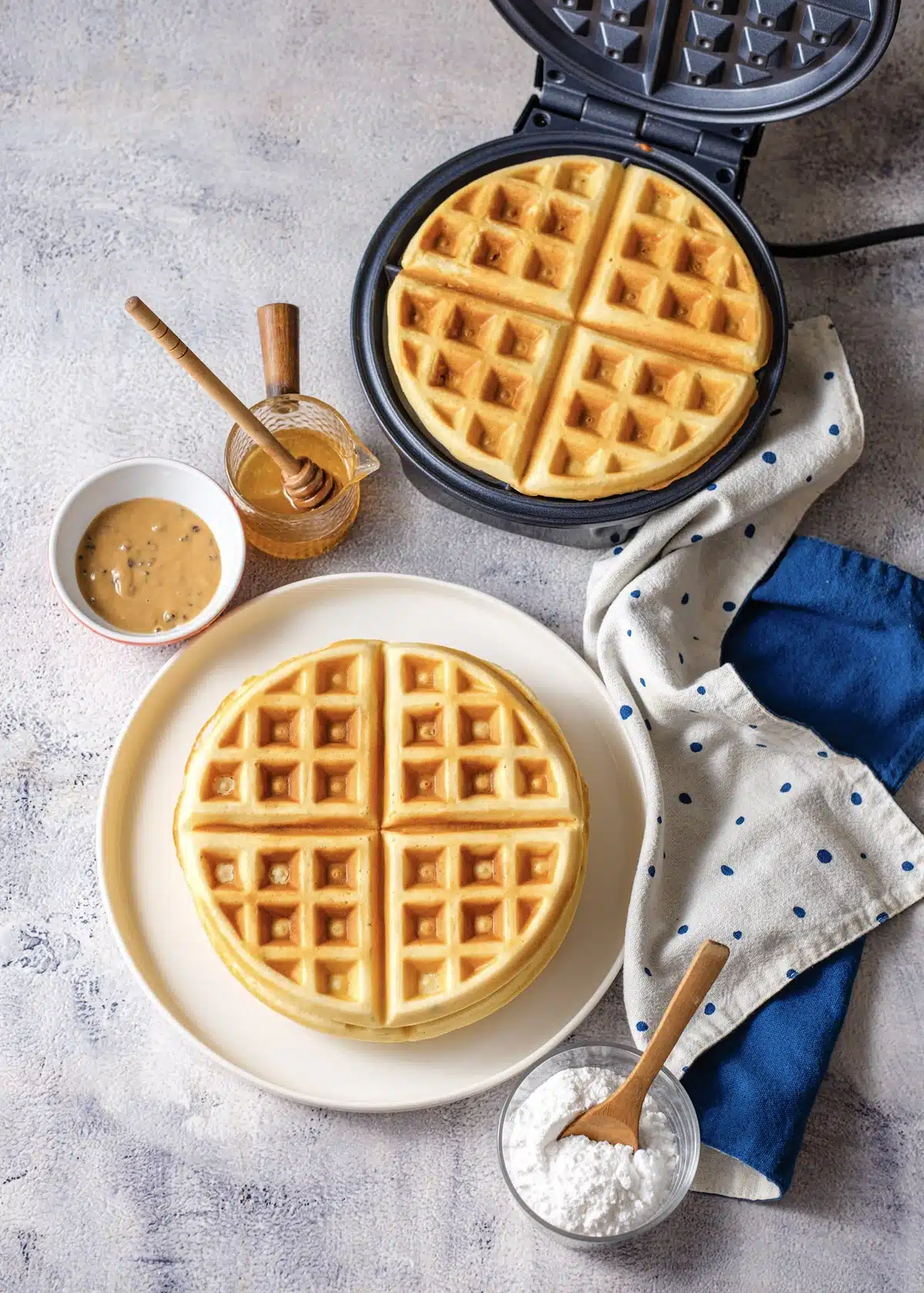 Bisquick waffles stacked on a white plate in front of a round waffle maker, with a honey dipper, a bowl of peanut butter sauce and powdered sugar