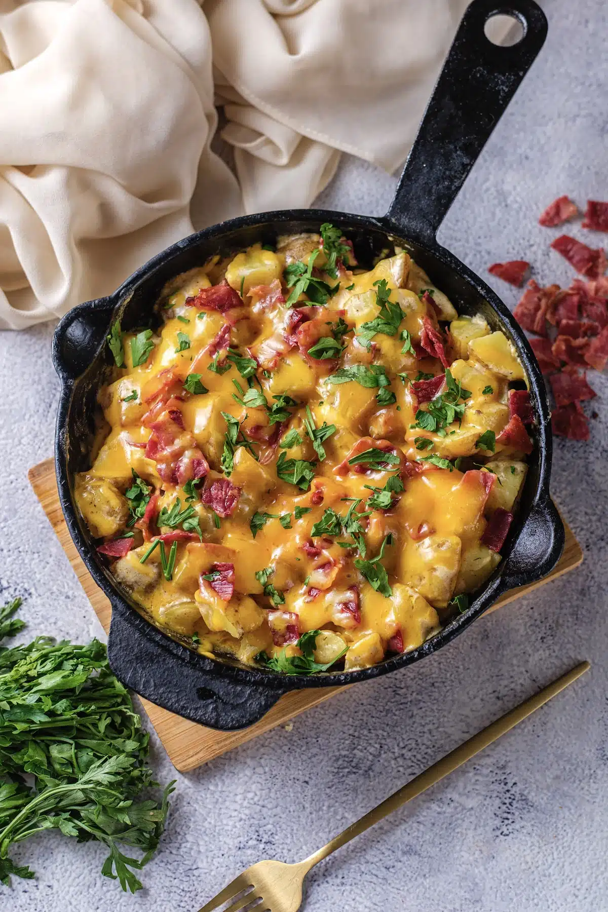 Cheesy ranch potatoes in a cast-iron skillet on a wooden board, topped with melted cheddar, chopped turkey bacon and parsley, seen from above