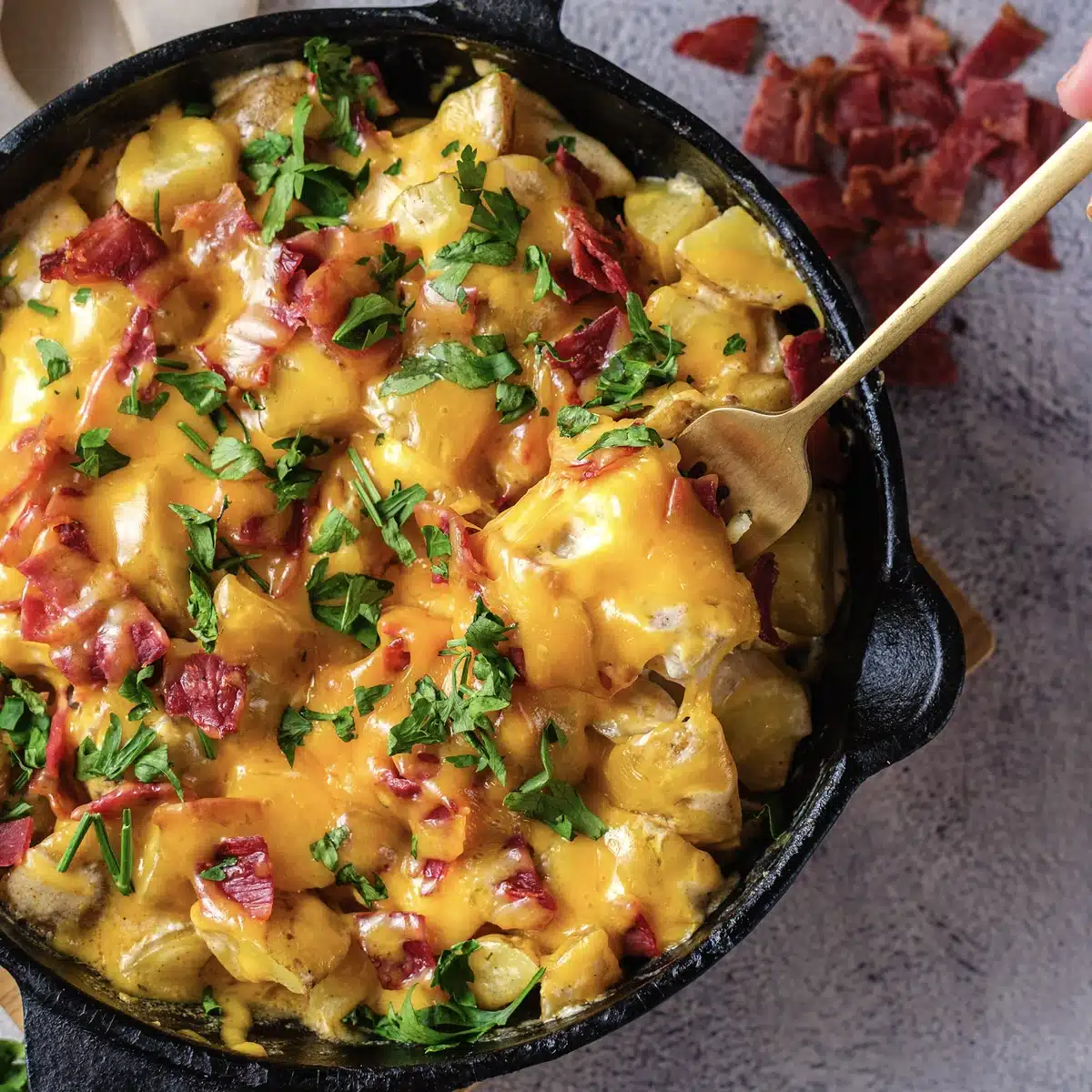 Cheesy ranch potatoes in the skillet with a gold spoon lifting a cheddar-covered chunk, seen from above