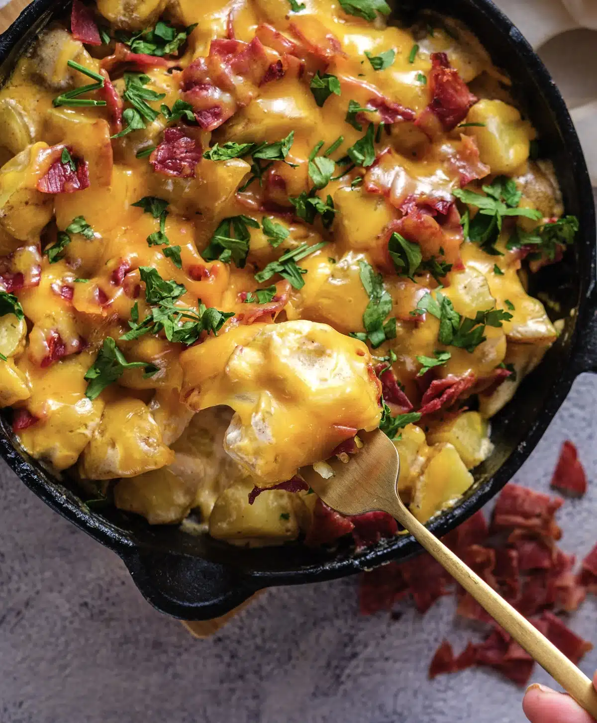 A gold spoon lifting golden chunks and melted cheddar out of a black skillet, with chopped cured meat scattered on the table