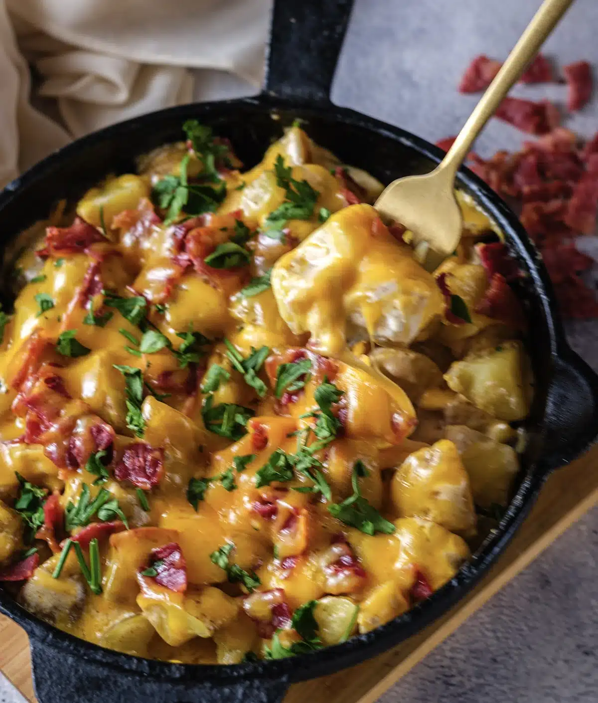 The pictured skillet of cheesy ranch potatoes with a gold spoon dug in, melted cheddar, chopped turkey bacon and parsley on top