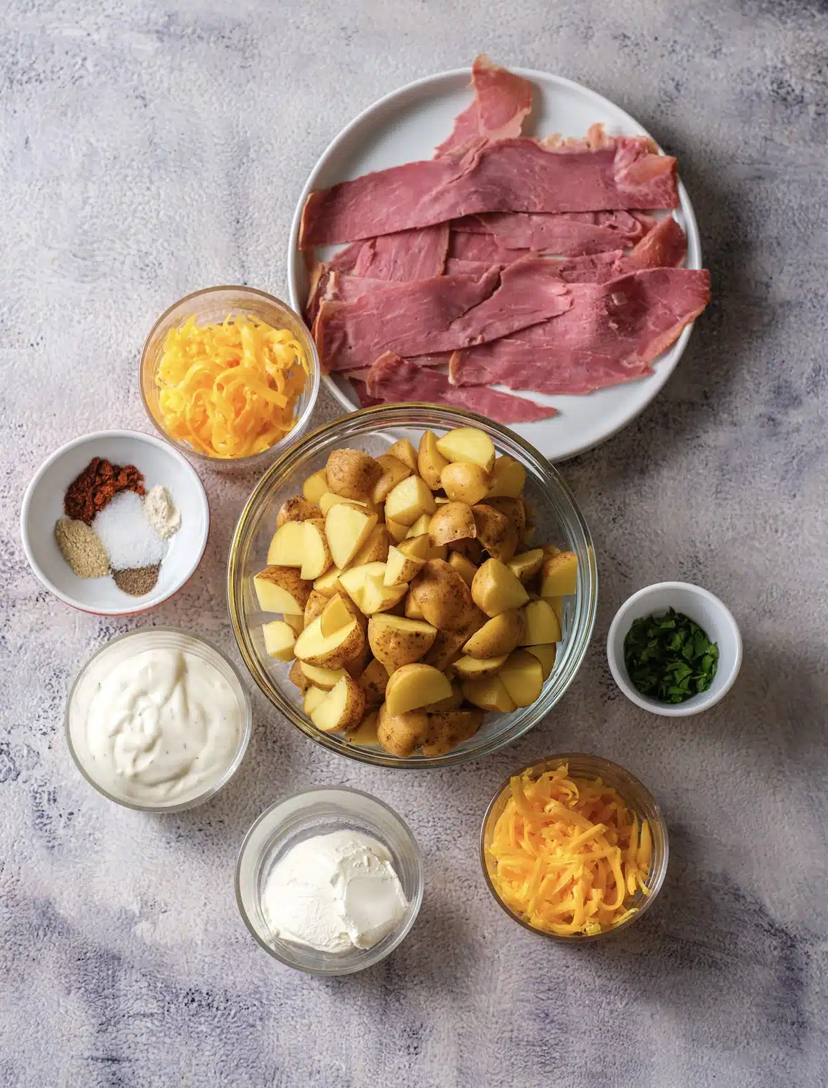 Ingredients for cheesy ranch potatoes: boiled potato chunks, shredded cheddar, sour cream, ranch dressing, spices, parsley and slices of turkey bacon