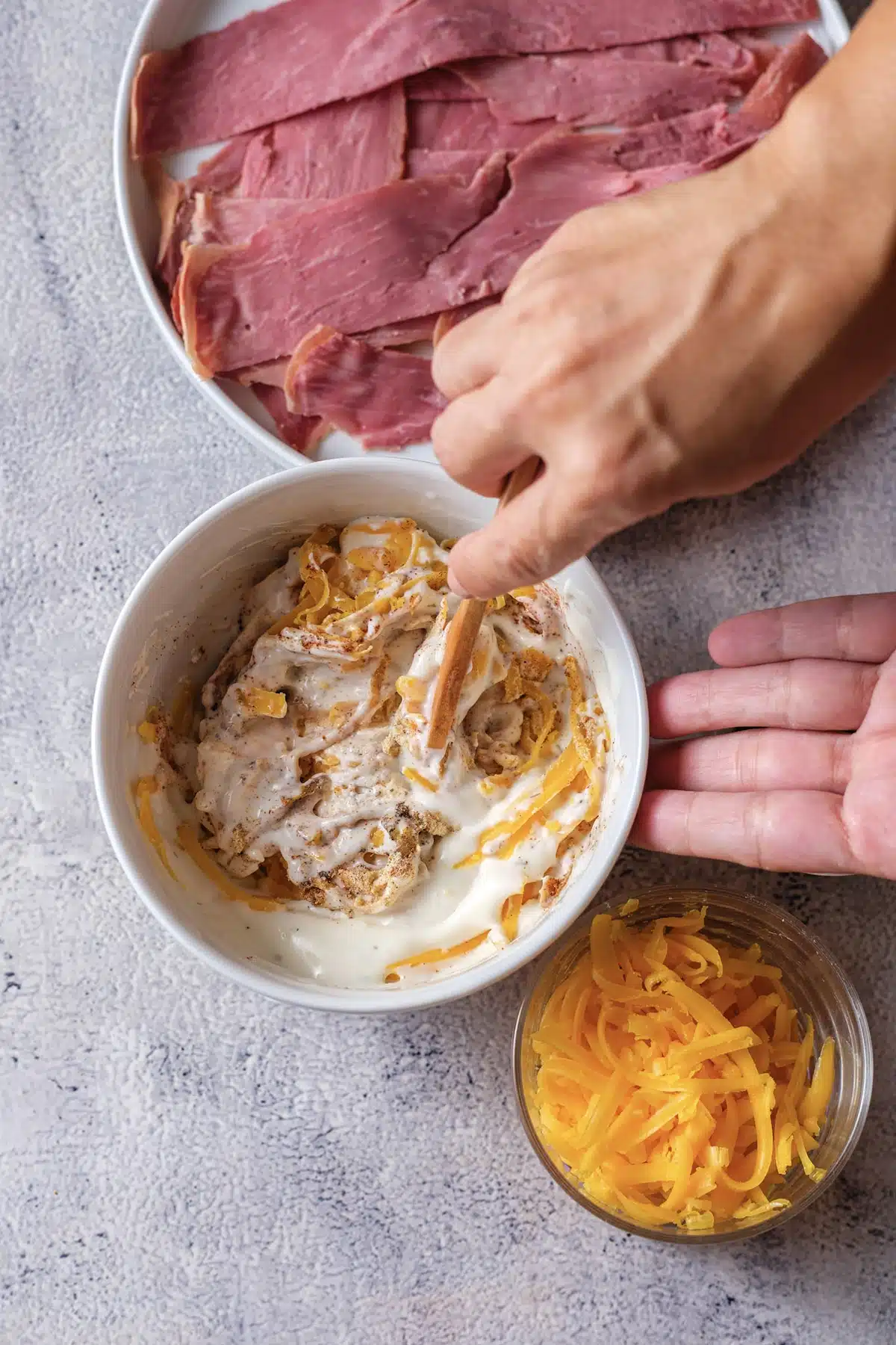 A hand stirring shredded cheddar and spices into a bowl of sour cream and dressing, with a dish of sliced cured meat behind