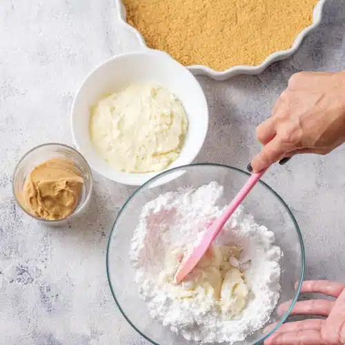 Powdered sugar and cream cheese beaten together in a glass bowl with a pink spatula, with the graham crust and a dish of creamy spread beside