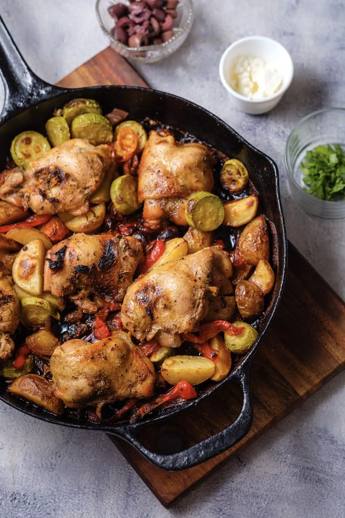 Greek sheet pan chicken made in a cast-iron skillet, with roasted thighs, potato wedges and zucchini, and bowls of olives, feta and parsley beside it