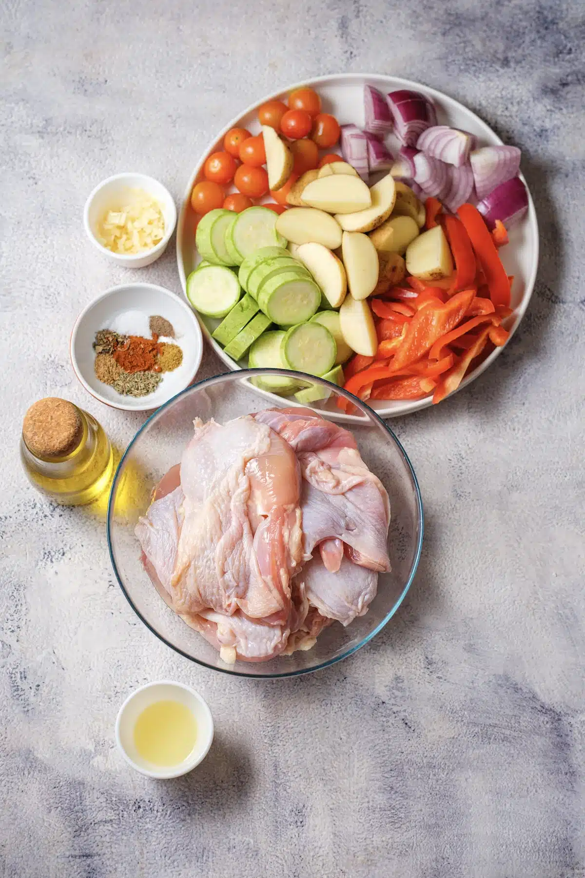 Ingredients for Greek sheet pan chicken: raw thighs, potato wedges, zucchini, red pepper, red onion, cherry tomatoes, olive oil, lemon juice, garlic and spices