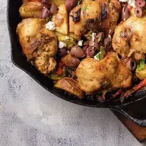 A serving of Greek sheet pan chicken: a roasted thigh with potatoes, zucchini, olives and feta in the skillet
