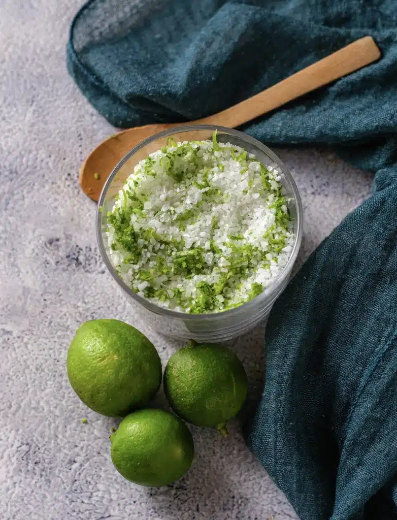 Lime salt in a glass jar with a wooden spoon and three limes on a blue cloth, seen from directly above