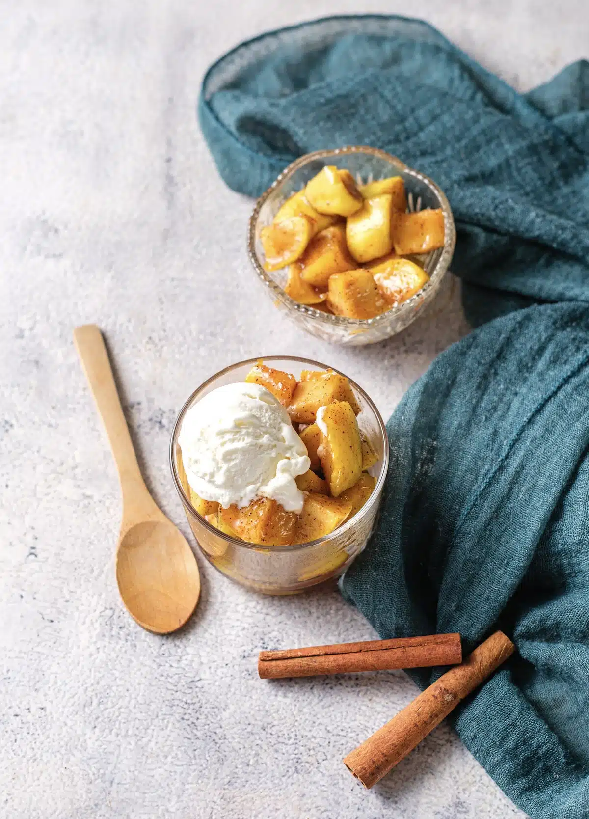 Microwave baked apples in a glass cup topped with vanilla ice cream, a second cup of the apples behind, with a wooden spoon and cinnamon sticks, seen from above