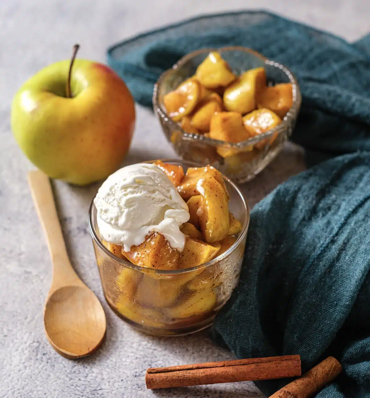 Microwave baked apples under a scoop of vanilla ice cream in a glass, a bowl of apples and a whole apple behind on a teal cloth