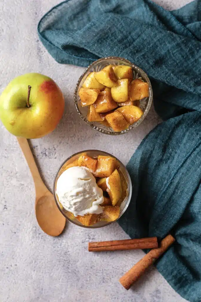 Microwave baked apples in a glass with vanilla ice cream and a second glass of cinnamon apples behind, with a whole apple and a wooden spoon, seen from directly above