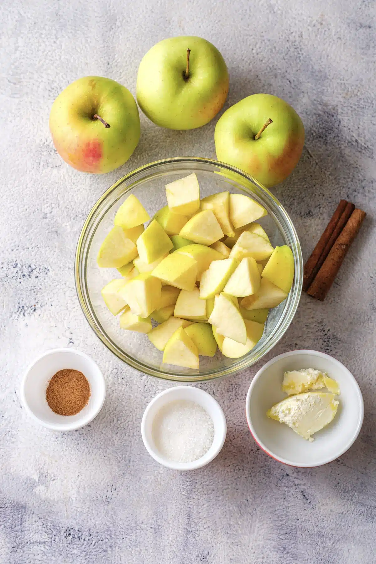 Ingredients for microwave baked apples: three apples, a glass bowl of apple chunks, and small dishes of cinnamon, sugar and butter with cinnamon sticks