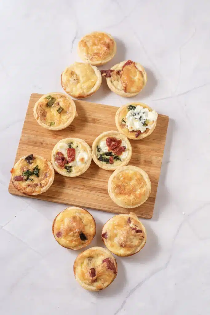 Mini quiche with four fillings arranged on a wooden board over a marble counter, seen from directly above