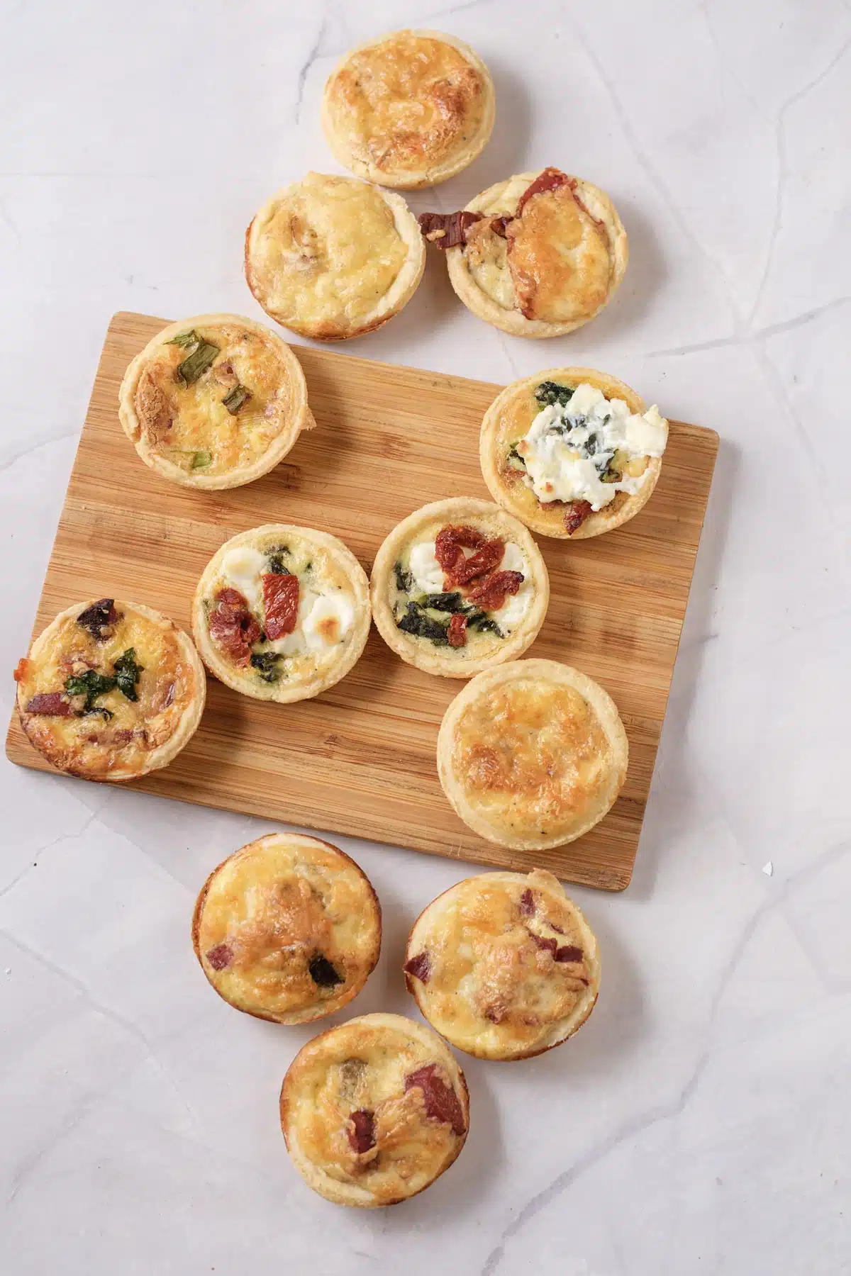 Twelve mini quiche with bacon, spinach, goat cheese and sun-dried tomato fillings on a wooden board and a marble counter, seen from above