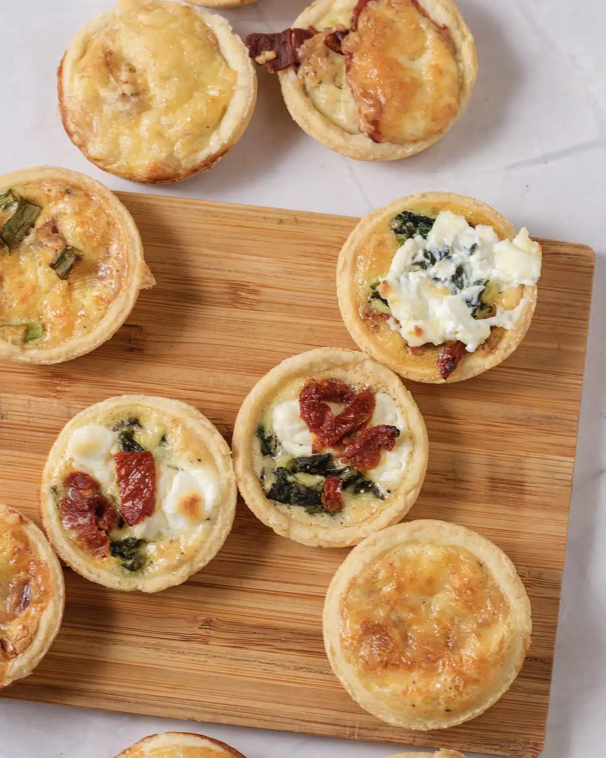 Close view of mini quiche on a wooden board: one topped with goat cheese and spinach, one with sun-dried tomato, one plain and golden