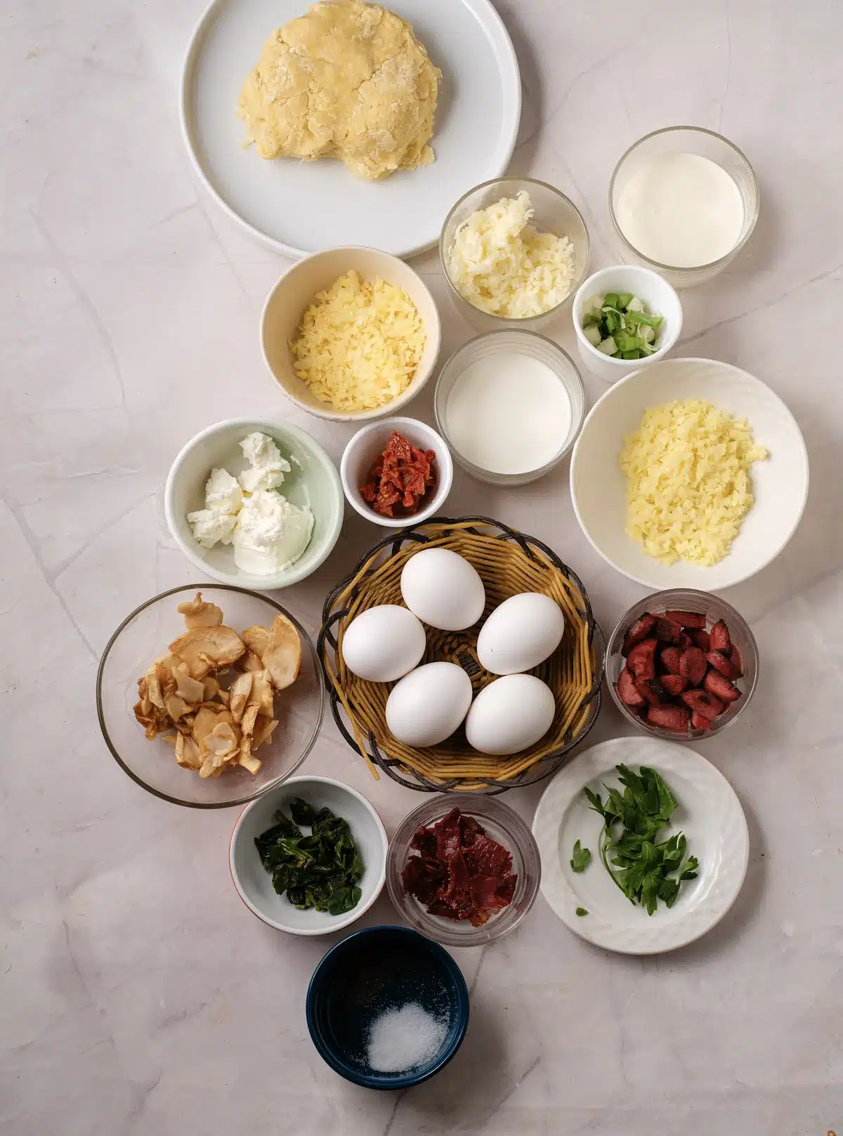 Ingredients for mini quiche: pie dough, shredded cheese, cream, milk, eggs, goat cheese, chopped ham, bacon, spinach, sun-dried tomatoes, green onions and salt in small bowls