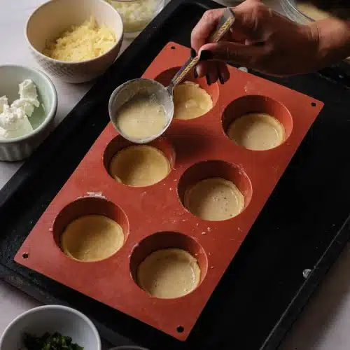 Custard ladled from a small pan into the first pastry-lined cup of a red silicone mold