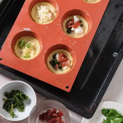 The filled cups topped with cheese, ham, spinach and sun-dried tomato, with dishes of fillings beside the mold