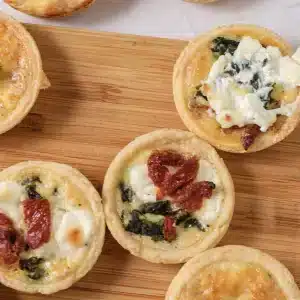 A single mini quiche topped with goat cheese and spinach on a wooden board, with others around it