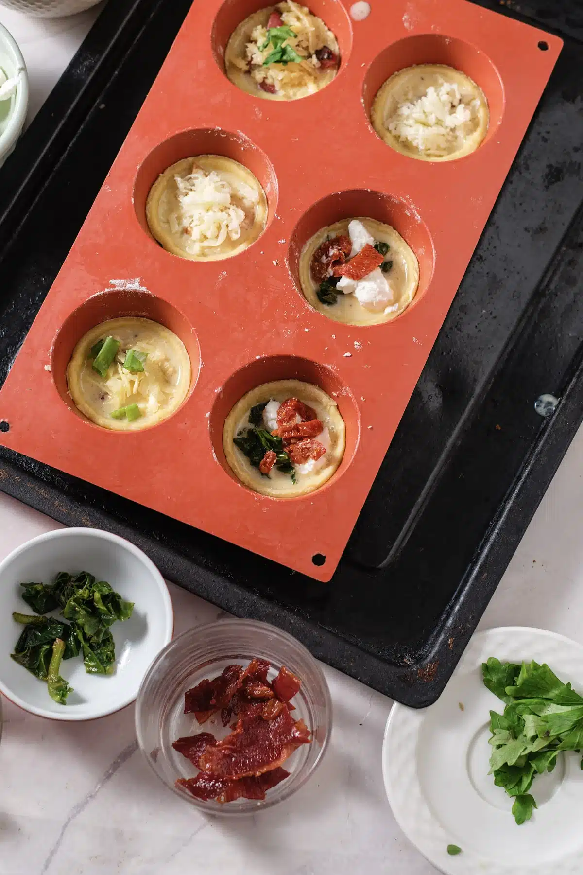 A red silicone mold on a black sheet pan with custard-filled pastry cups topped with cheese, spinach, ham and sun-dried tomato, ready for the oven