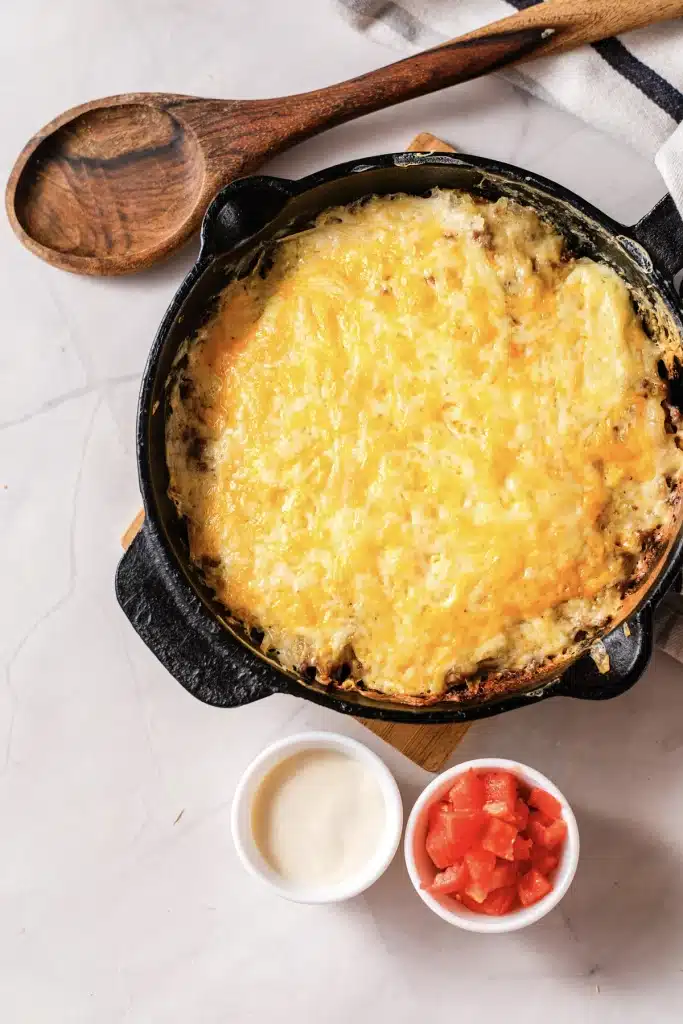 Taco hashbrown casserole in a cast-iron skillet with a wooden spoon, sour cream and diced tomato, seen from directly above