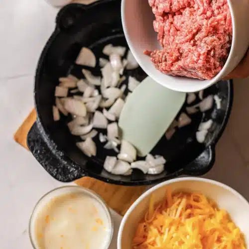 Raw ground beef tipped from a bowl onto the onion in the skillet