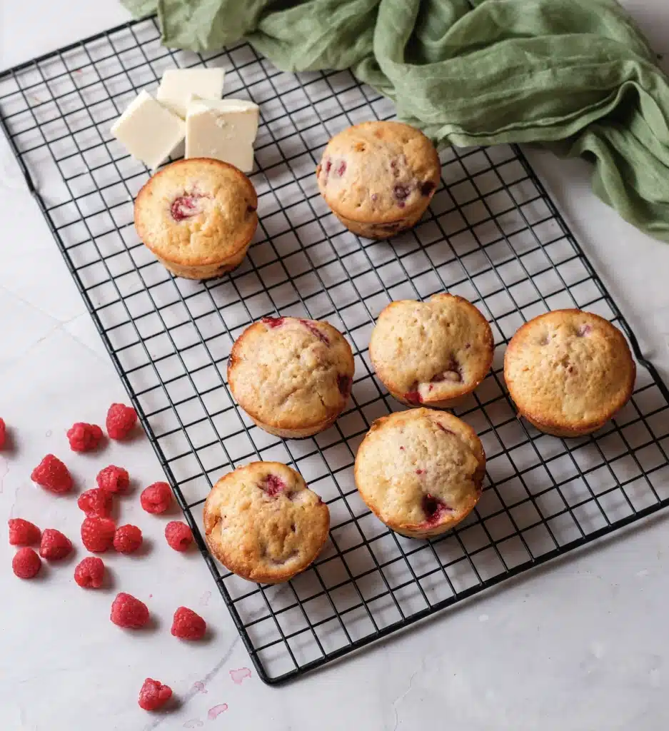 White chocolate raspberry muffins on a black wire rack with fresh raspberries, blocks of white chocolate and a green cloth, seen from directly above