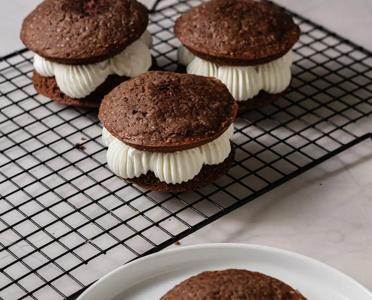 Three old-fashioned whoopie pies with piped vanilla filling on a wire rack beside a white plate