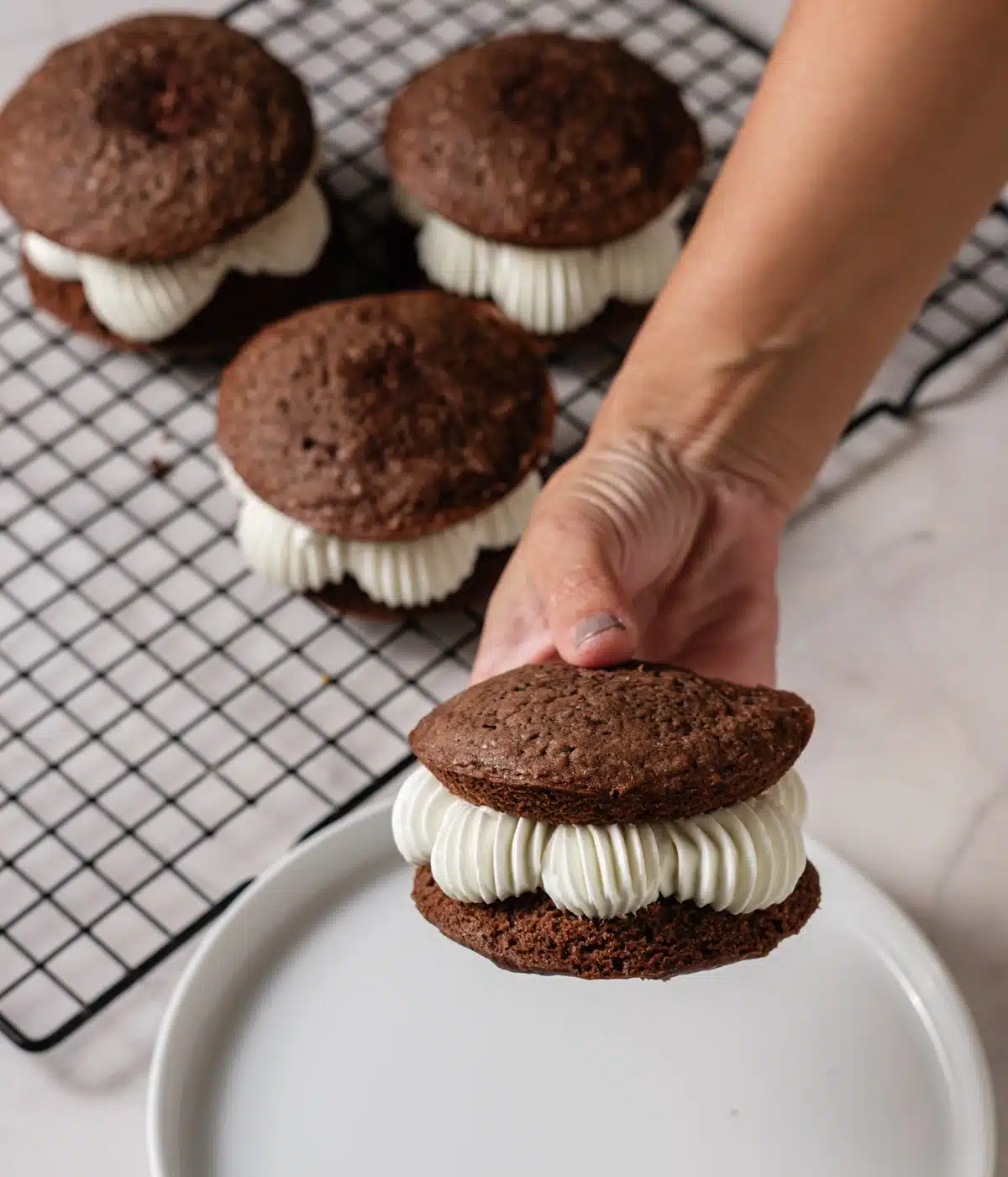 A hand lifting a filled whoopie pie from a wire rack of chocolate cakes with piped cream