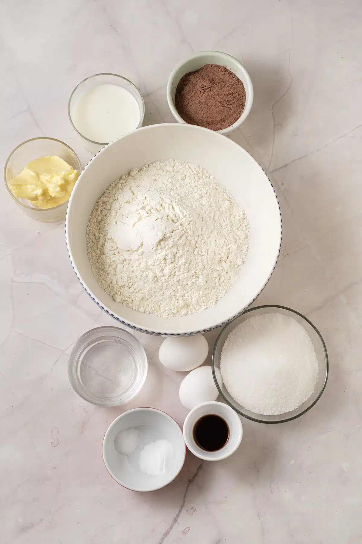 Ingredients for whoopie pies: flour, cocoa, sugar, buttermilk, butter, an egg, vanilla, baking soda and hot water laid out on marble
