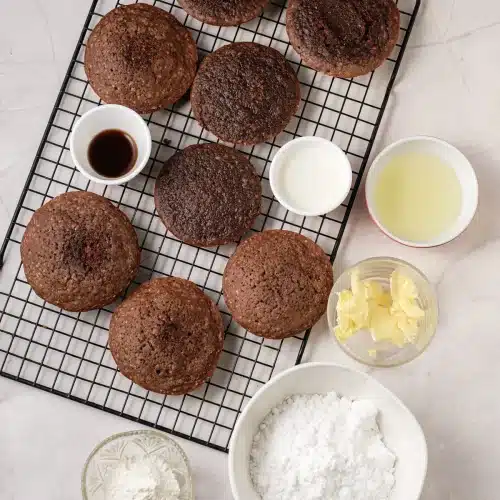 Baked cocoa cakes cooling on a black wire rack with bowls of powdered sugar, butter, milk and vanilla beside them, seen from above