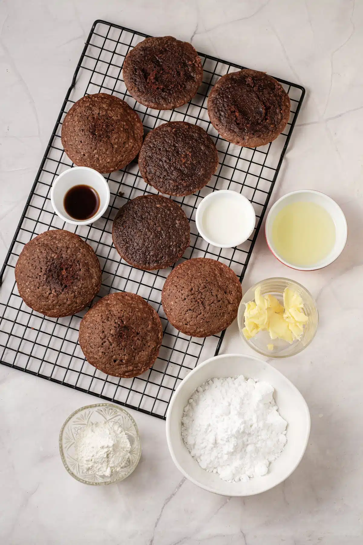 Baked cocoa cakes cooling on a black wire rack with bowls of powdered sugar, butter, milk and vanilla beside them, seen from above